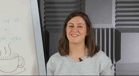 A white woman with shoulder length brown hair stands in front of a whiteboard, smiles and shrugs