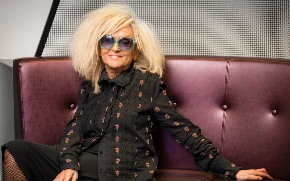 A photo of Annie Nightingale smiling towards the camera and sitting on a couch.