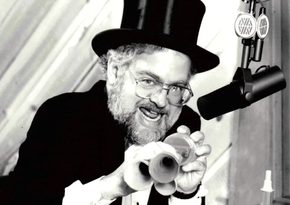 A black and white photo of Dr. Demento, wearing a top hat and holding an air horn, behind a micrphone.