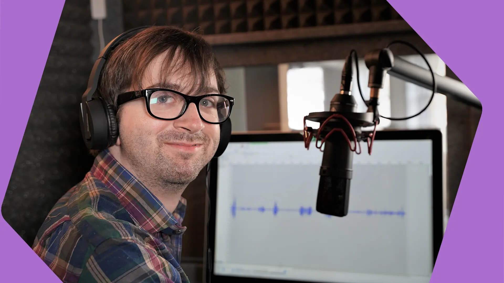 A white man with dark hair and black glasses wears headphones and smiles at the camera. He is sat in a studio in front of a microphone and a screen with sound levels on it in white. He is wearing a plaid shirt. The borders to this image are purple.