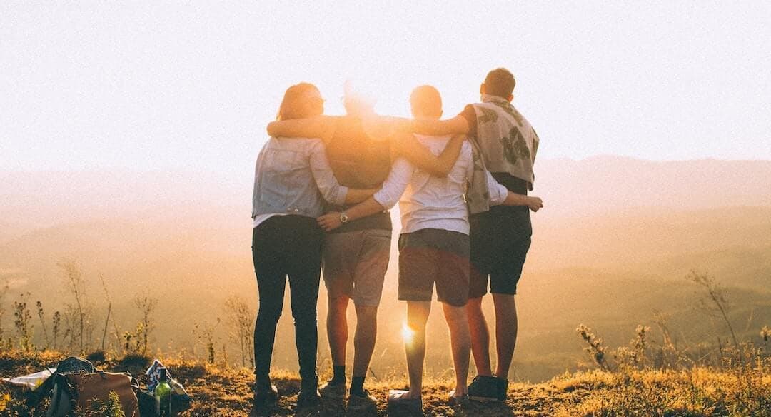 This is an image of four friends with their arms round one another. They are stood on a hill looking out to a sunset, and the sunset covers their features so its hard to tell who is who or what they look like as they are silhouetted.
