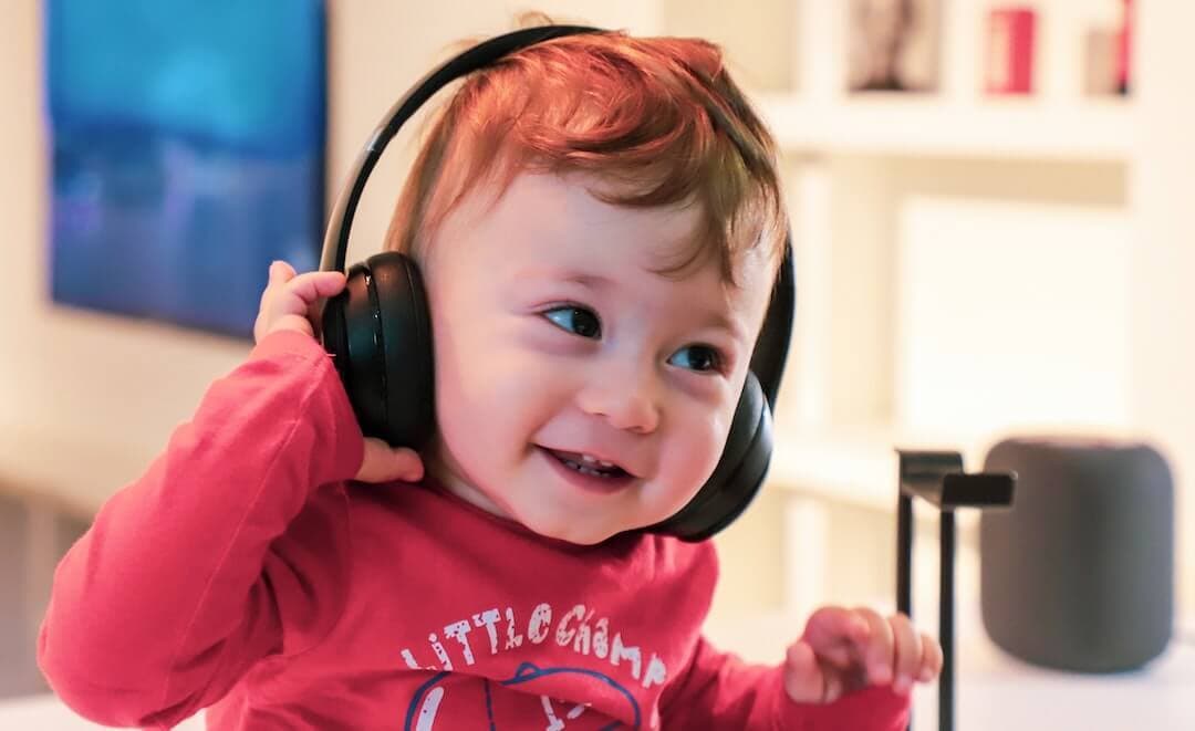 This is an image of a baby with headphones on. The baby is a white boy with brown hair. He is wearing a red hoodie and has one hand on the headphones. The background is a neutral white.