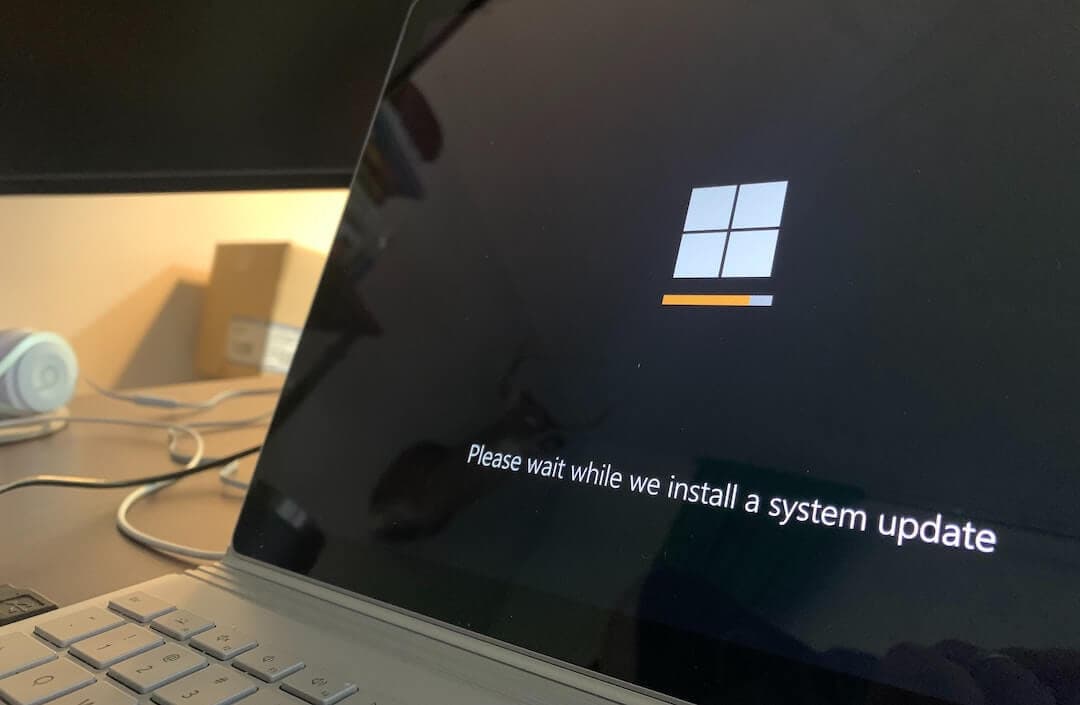 This is a zoomed in image of a Surface laptop. In the centre of the desktop is a white Windows square and an orange loading bar. It reads "Please wait while we install a system update". The background is completely black.