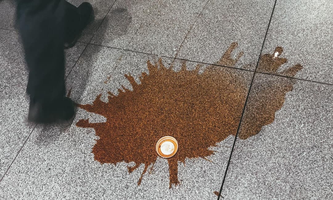 This is an image of spilt coffee. There is a big brown pool of liquid on a marbled granite floor. There is the plastic white lid of a coffee in the centre of the image. On the right is a man in black trousers and plain black shoes walking.