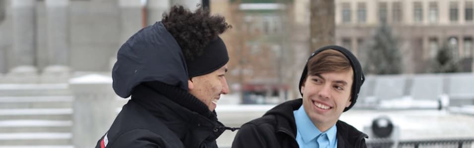 On the left a person with black hair speaks to a white amn with brown hair and a black hat on. Both are wearing winter clothes and appear to be walking in the street, against a city backdrop.