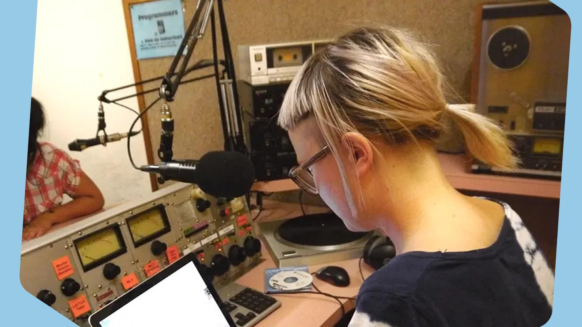 This is an image of a girl from behind working at a radio station. She is in front of a microphone, using a mixing desk. She wears glasses and a black t shirt.