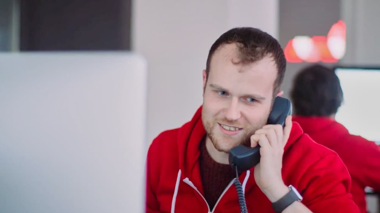 This is a picture of a white man with gingery brown hair on the phone. He is smiling and talking into it. The backdrop is white and neutral (though it shows someone in a red hoodie with their back to the image) and the man is sat in front of a Mac.