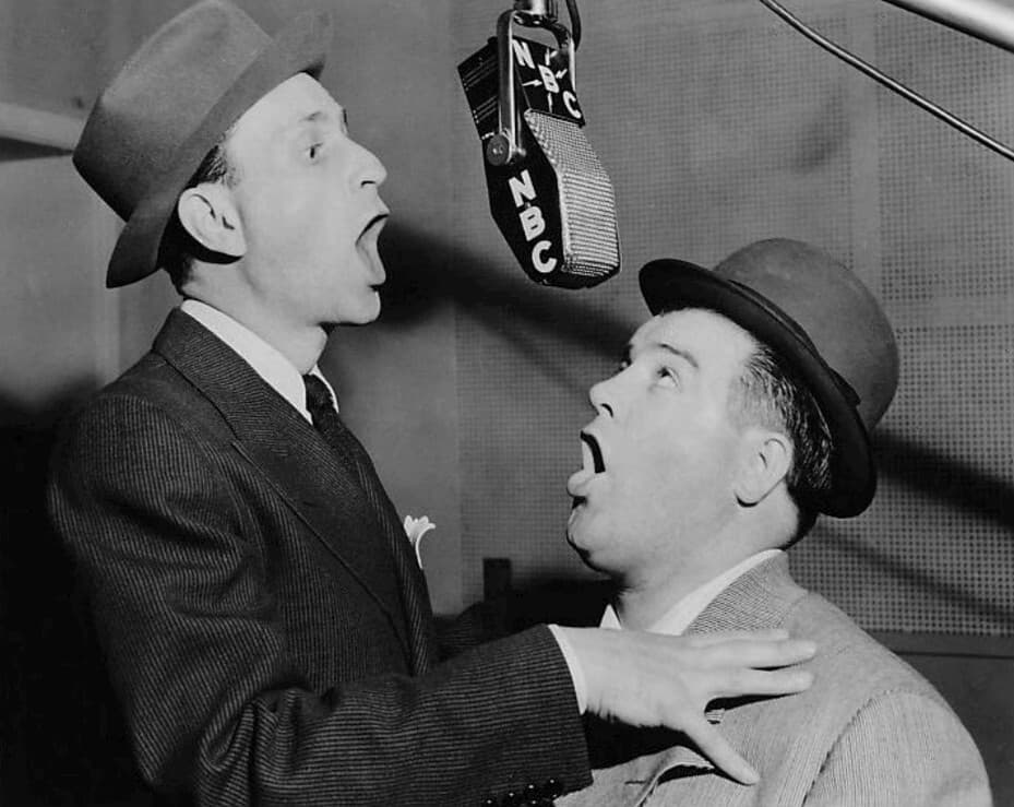 A black and white photo of Abbott and Costello singing in to a microphone.