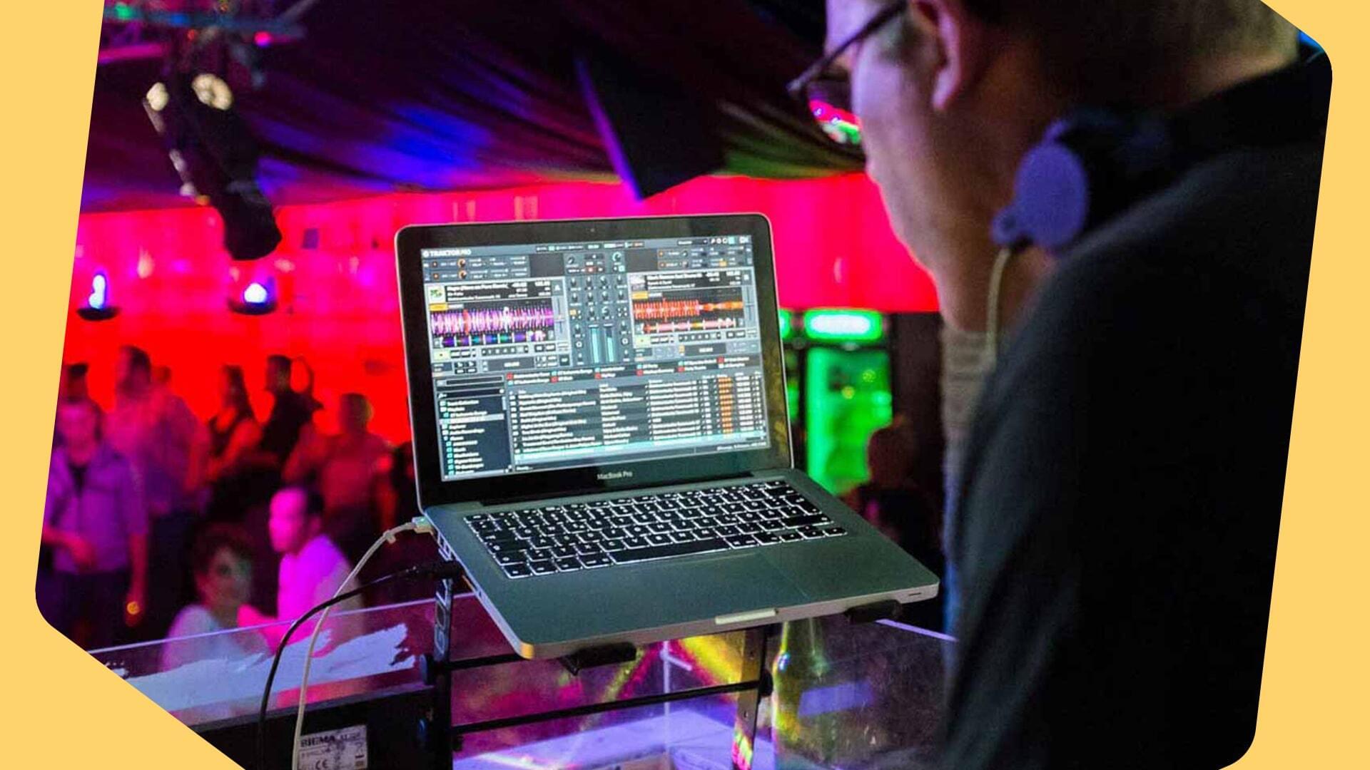 This is an image of a red and pink club or event with an audience visible. In the forefront of the image is a Macbook open on audio editing software with the back of a person leaning towards it. The image has yellow borders.