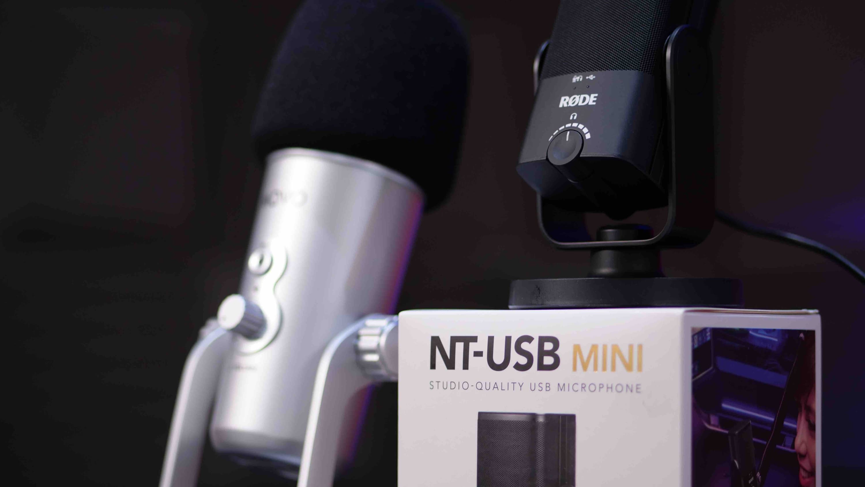 Image shows the RODE NT-USB Mini mic placed upon its box, with a silver blue yeti microphone in the background.