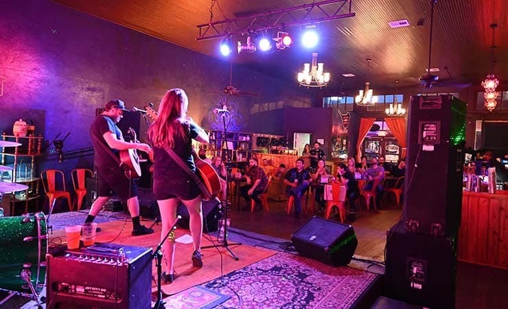 Image shows one male and one female guitarists playing to a seated audience in a bar.