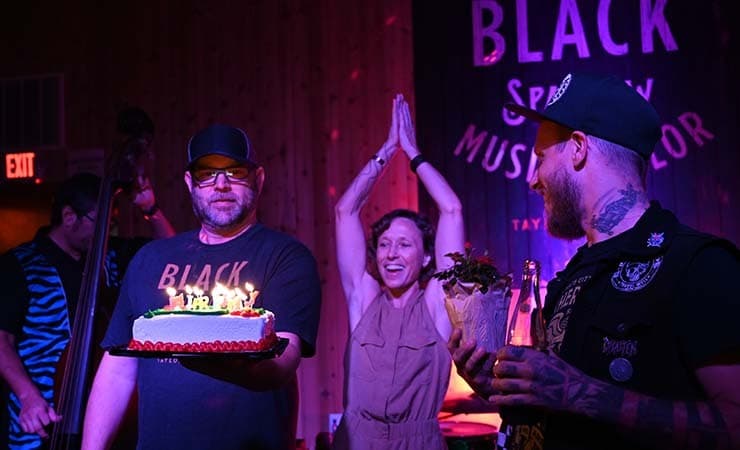 Image shows a man holding a candle-lit birthday cake, with a man and a woman cheering beside him.
