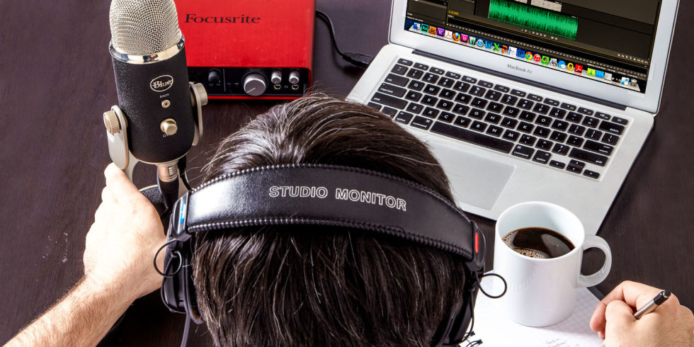 This is an overhead image of a person with short dark hear wearing headphones that read Studio Monitor. On the right is a cup of coffee in a white mug. On the left is a microphone and red audio interface. They are working at a macbook.