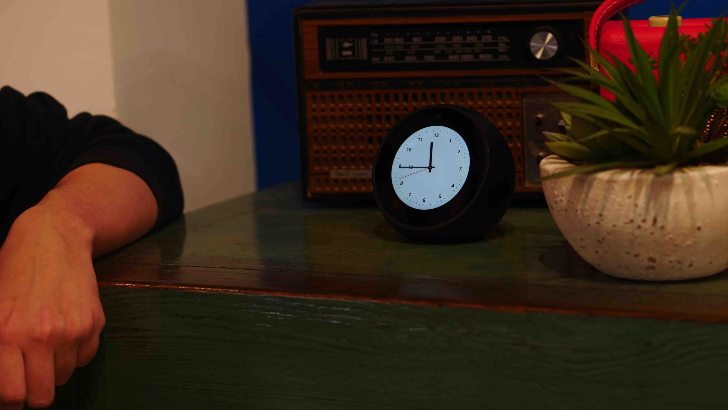 Image shows a small light-up clock on a chest of drawers, and someone's arm leaning against the chest.