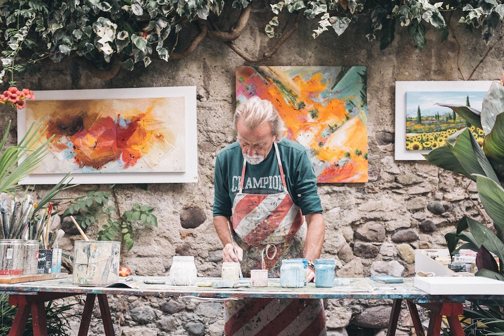 This is a man with grey long hair and a grey beard wearing a white and red striped apron and green shirt. He is painting something. The table in front of him is filled with art supplies, and behind him is a walled backdrop with framed art prints visible.