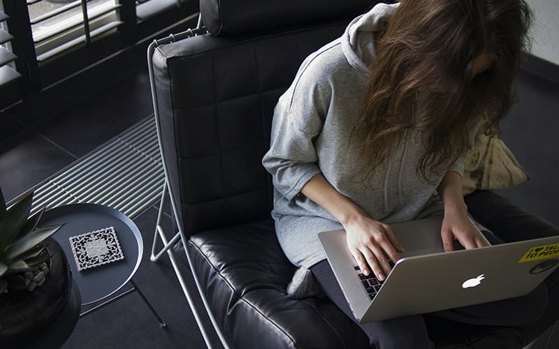 Image shows a woman on a Macbook, working remotely from a leather armchair.