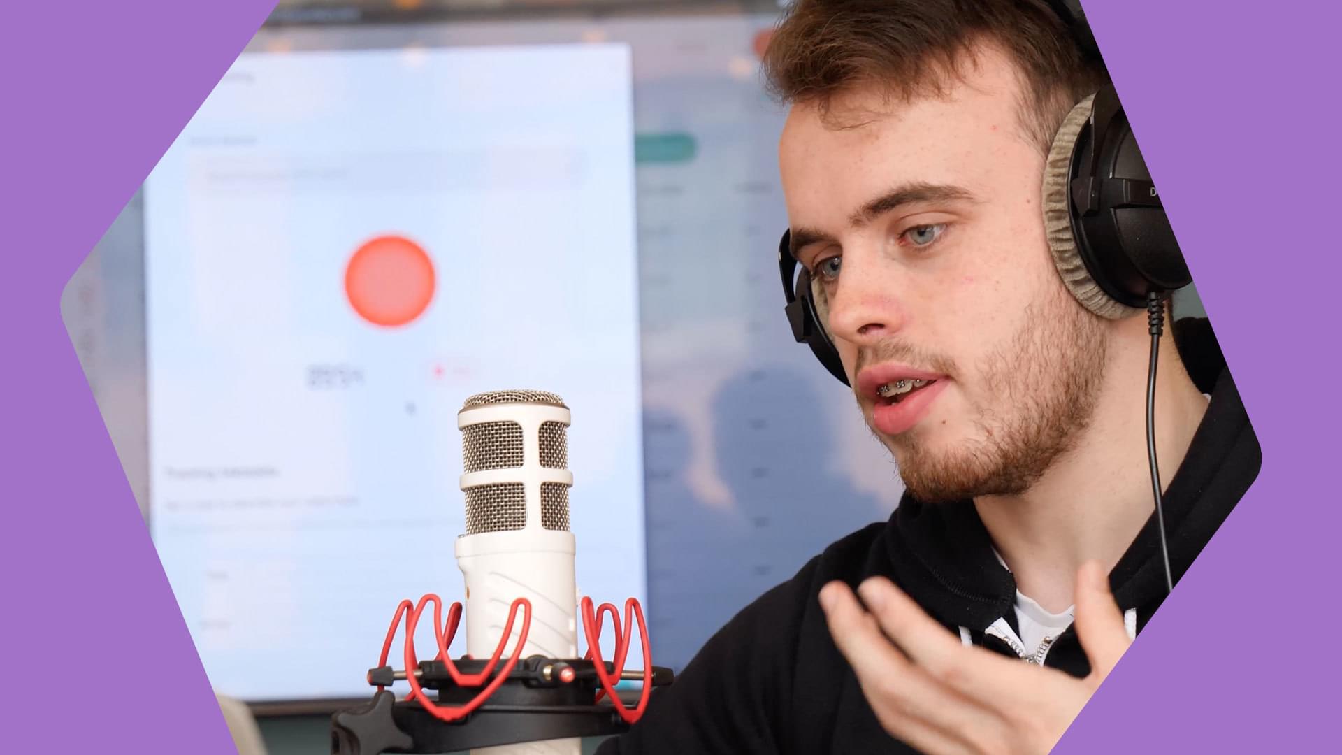 This is a white man with brown hair and a brown closely shaven heard. He is talking into a white and red microphone. The background is neutral but appears to be a powerpoint. The borders of this image are purple.