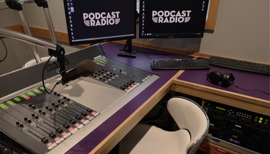 Image shows the inside of the Podcast Radio studio, with 2 broadcasting desks, 2 computer screens and a microphone on a desk.