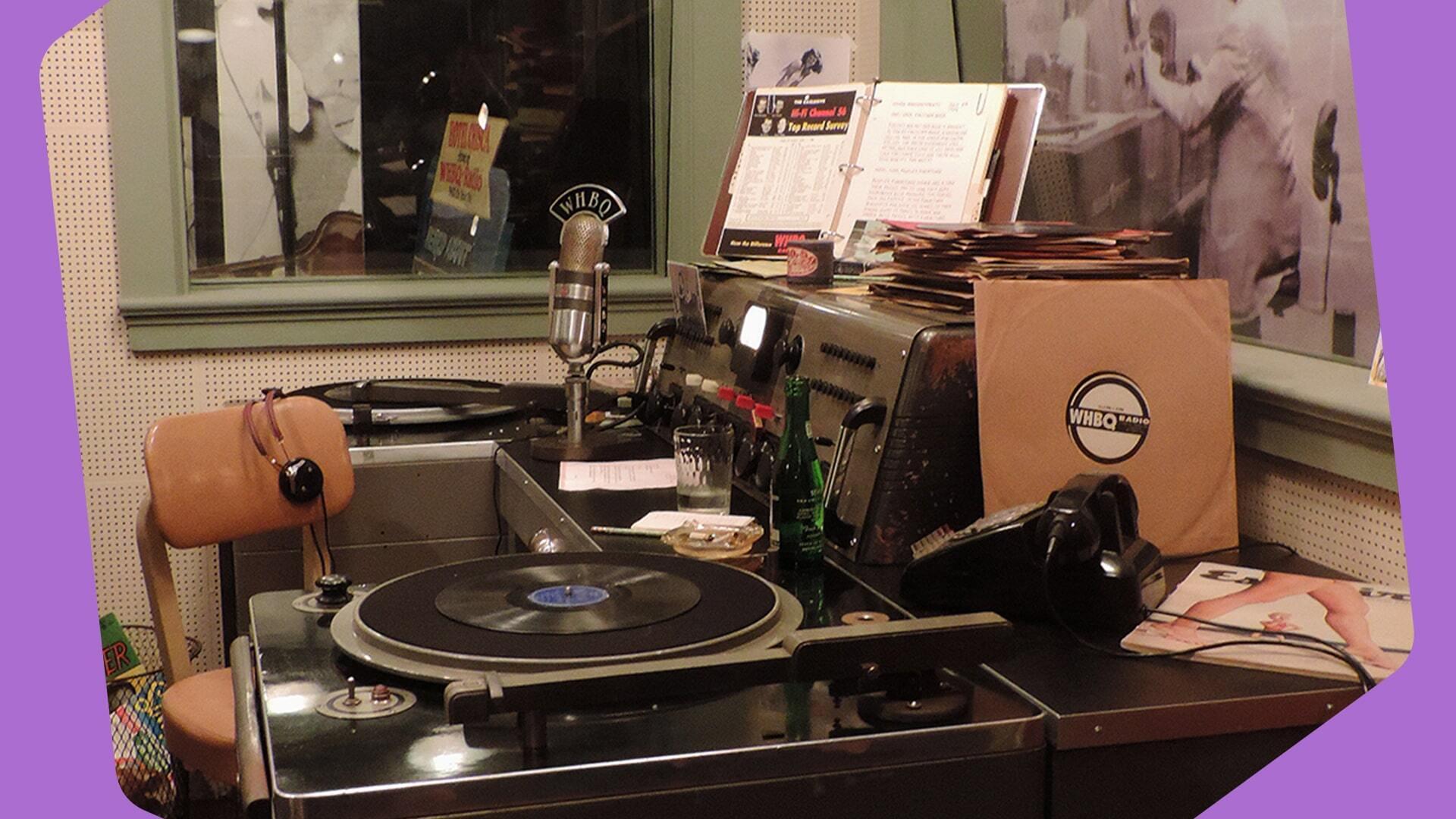 This is an image of a small makeshift studio setup. The corners of the image are purple. On the right there are some records, on the left there is a mixing desk and all over there are various small pieces of audio equipment.