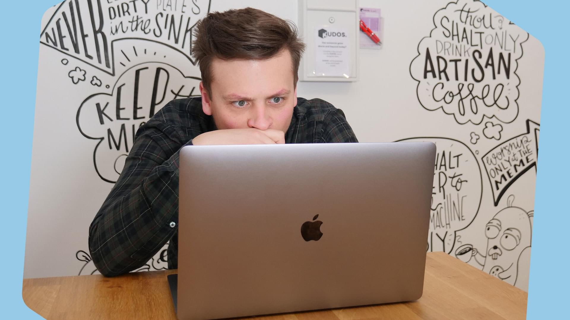 A white man with brown hair and blue eyes sits in front of a Macbook. He is reading and his expression is that of hyperfocus. The background is a white wall with patterned scribbles on it.