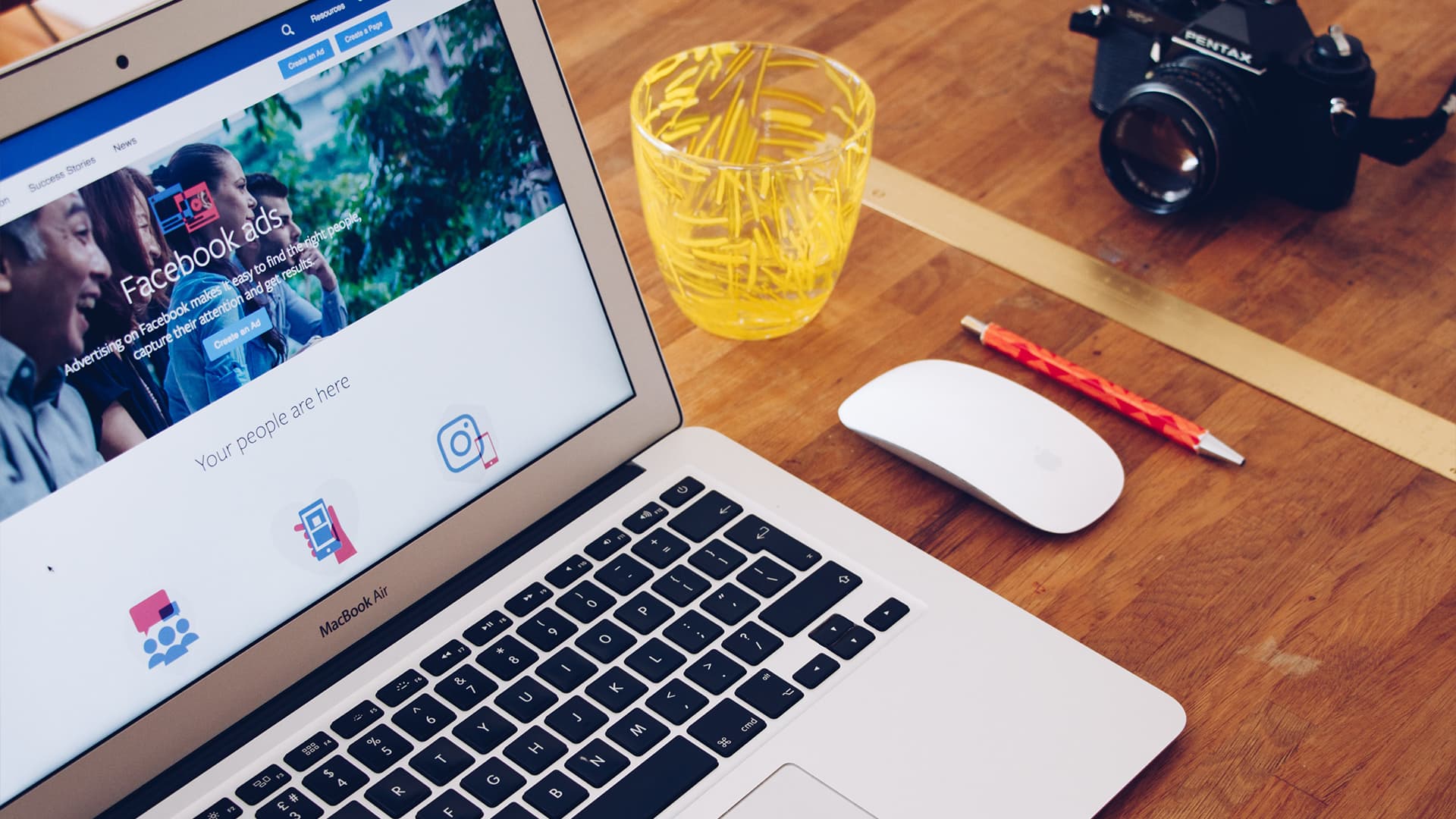 Image shows a Macbook, mouse, glass of water, pen and camera all neatly placed on a desk.