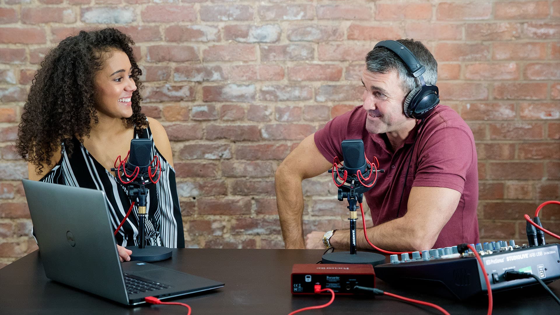 Image shows a male and female radio host, broadcasting with a laptop, a mixer and 2 microphones.