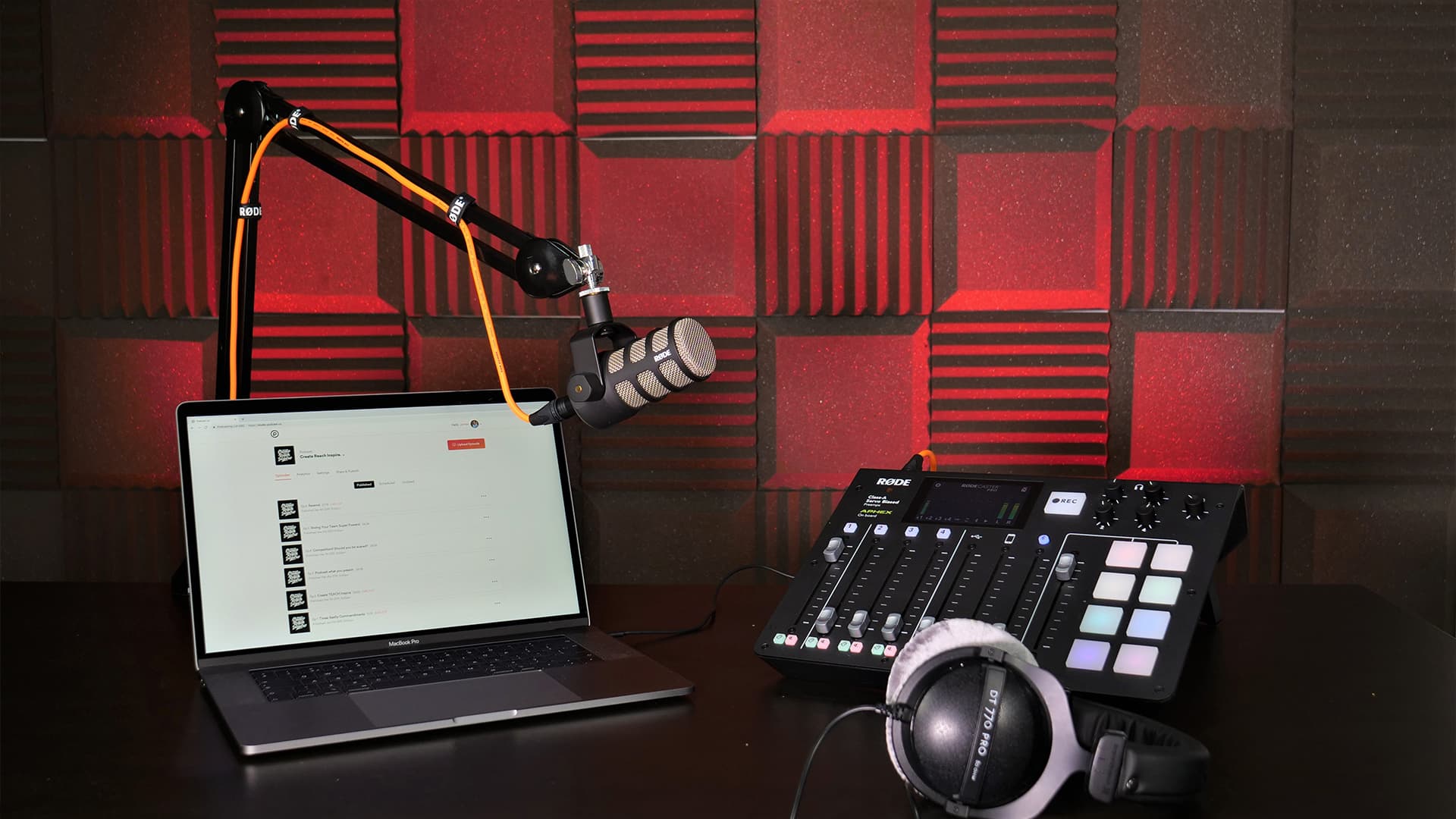 Image shows an online broadcasting set up: On a desk there is a laptop, a microphone in a stand, a Rodecaster mixer and a set of headphones.