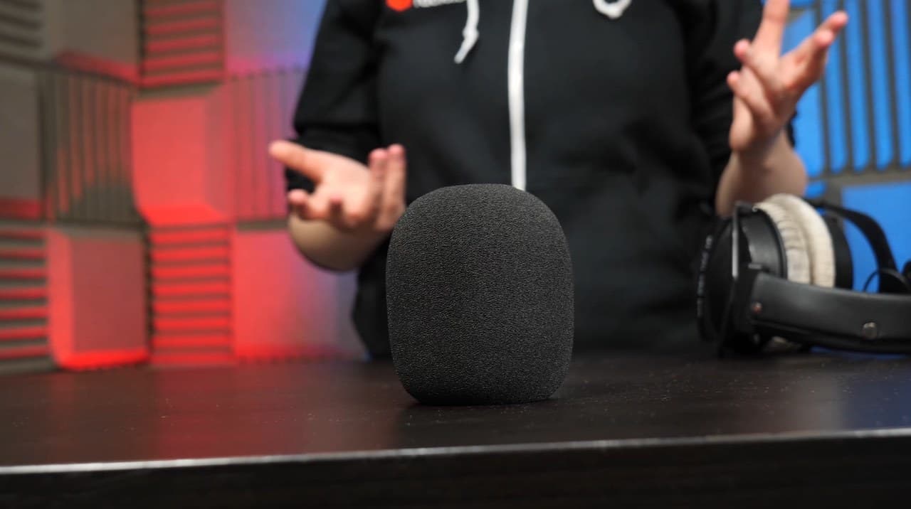 Image shows a black microphone windshield on a desk. There is a person and a pair of headphones in the background.