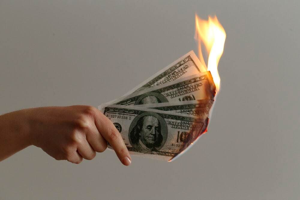 Image shows a hand holding some US dollar bills which are on fire.
