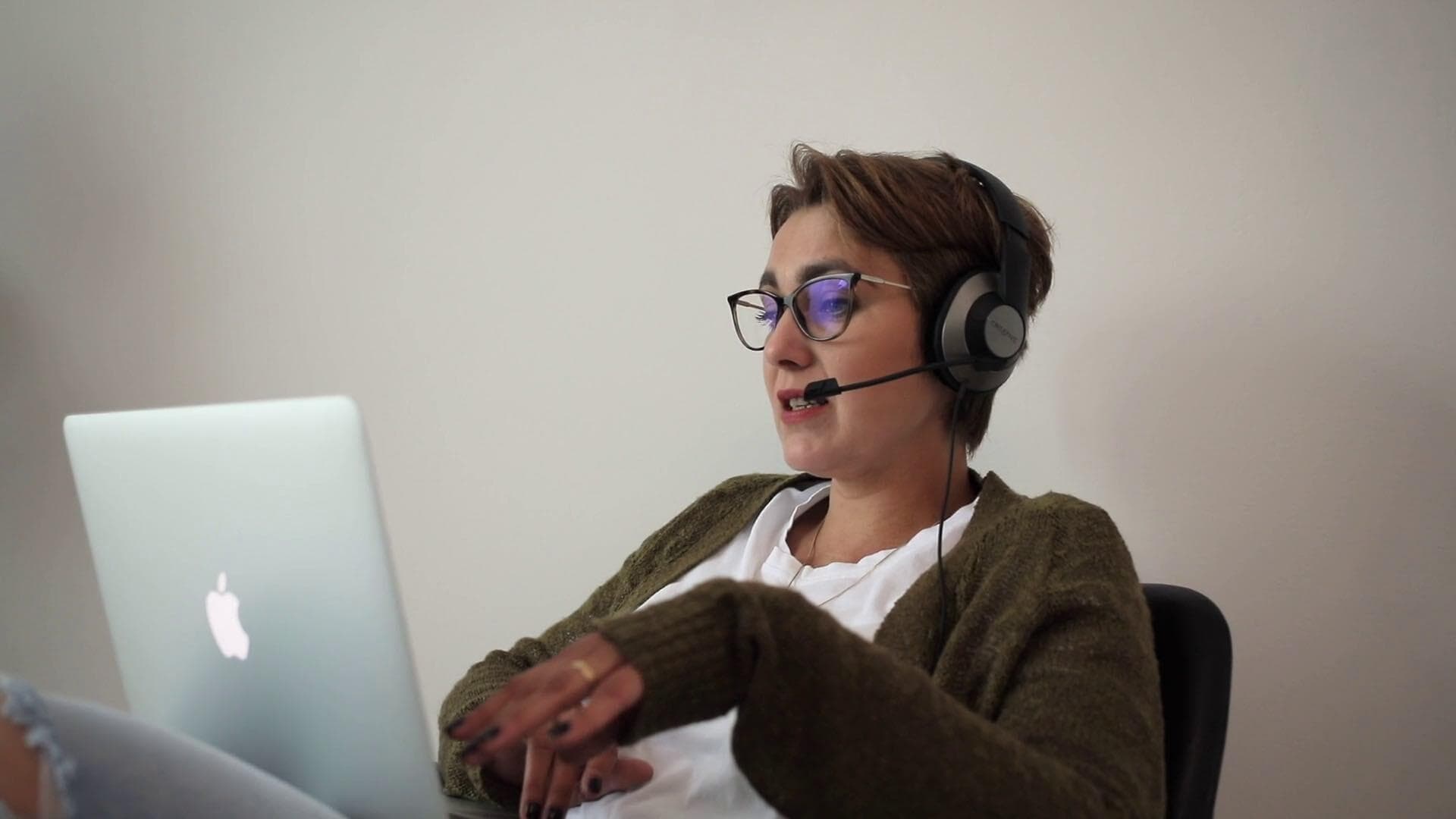 Image shows a young woman apparently on a Zoom call. She's has a Macbook resting on her lap and is talking into a headset.