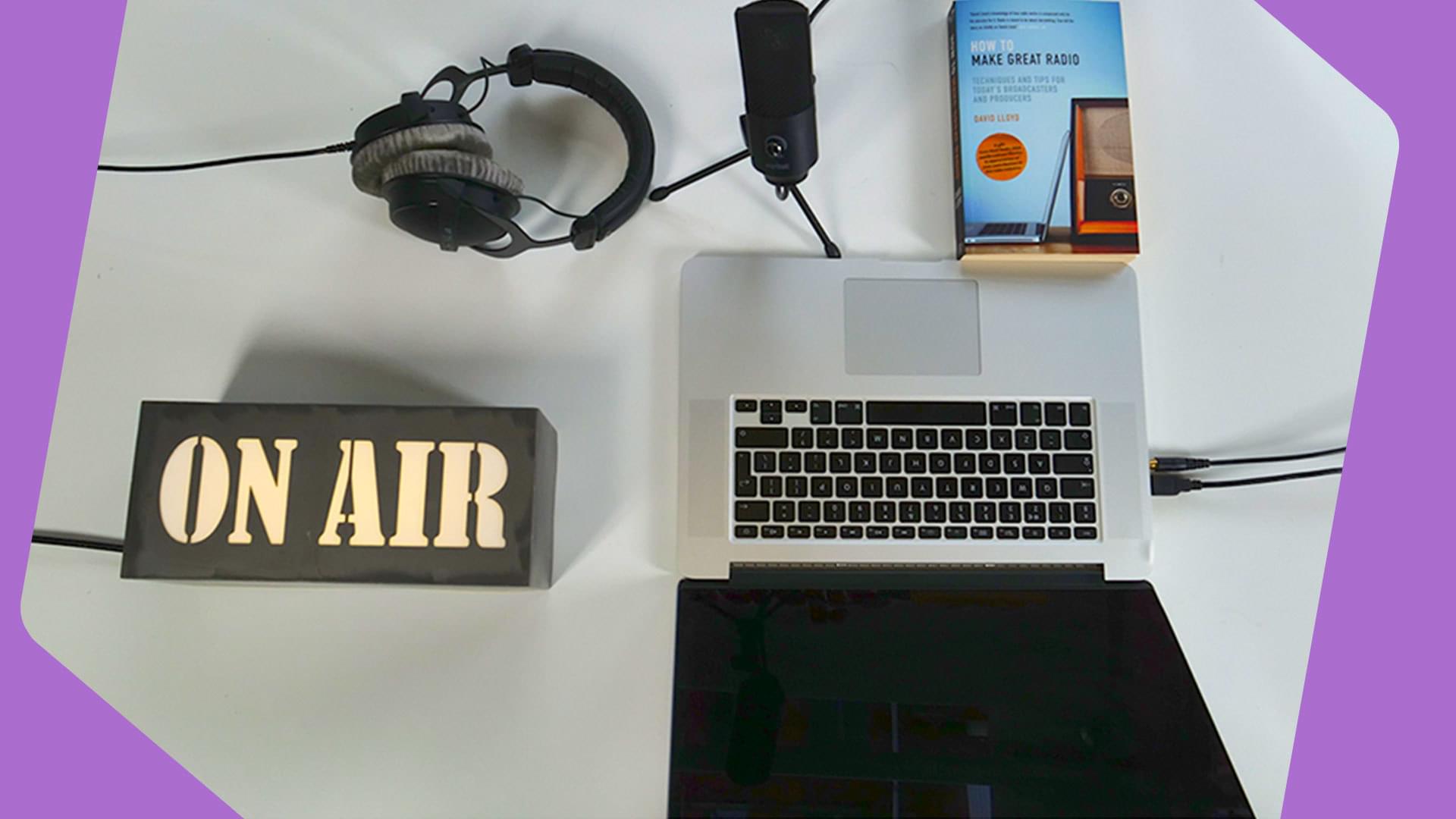 This is an image of an ON AIR black and white lightbox, an open Macbook, a pair of over-ear headphones, a Microphone and a book with an indistinguishable cover. There is a purple jaunty border to the image.