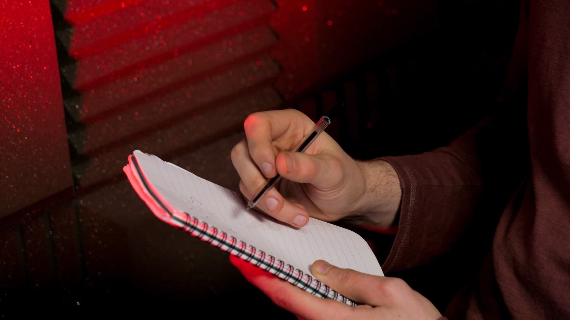 This is a neutral dark red background featuring a close up of white hands writing ideas for a radio script on a notepad. The pad is white with spiraled sides.
