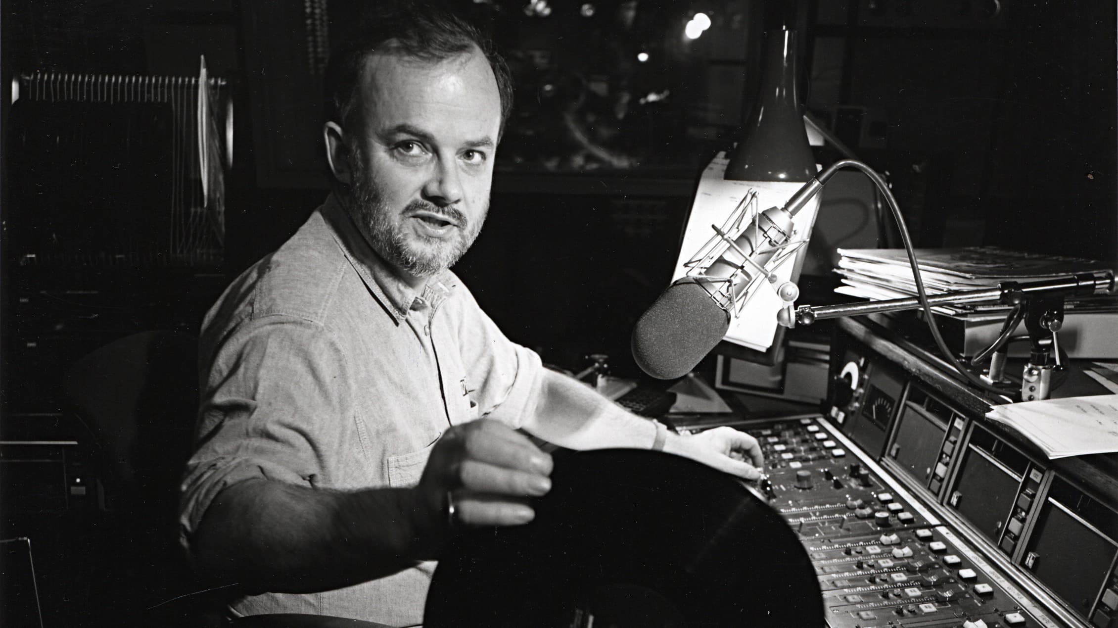 Image is a black and white photo of a young John Peel inside a radio studio.