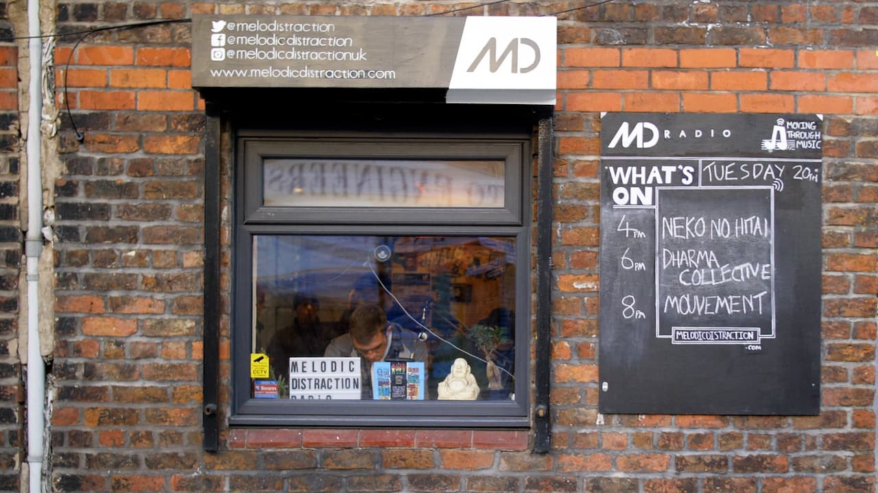 Image shows the outside of Melodic Distraction radio, there is a window looking into the station and a chalkboard on the wall which lists the schedule.