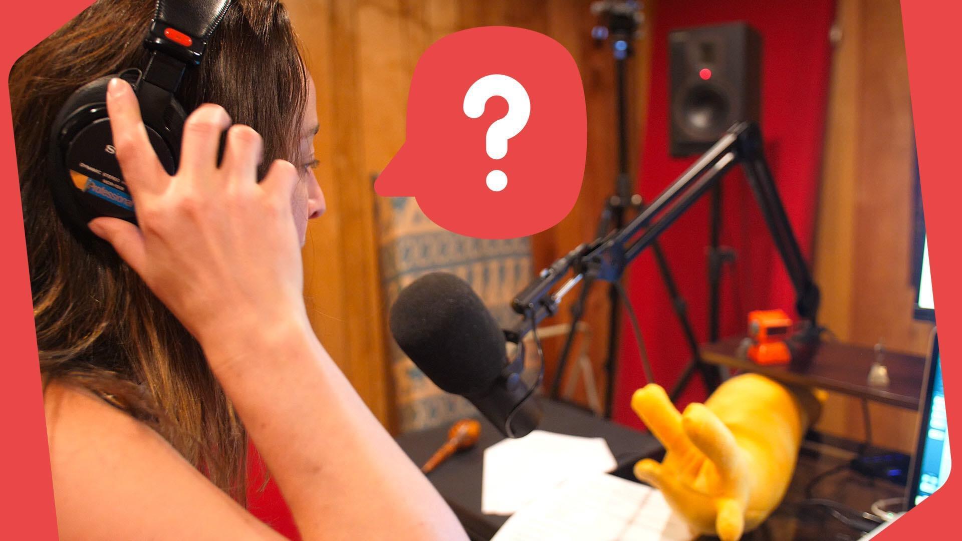 An image of a white woman with brown hair holding her hands to her headphones as she speaks into a microphone on a long stand. The background is a plain studio.