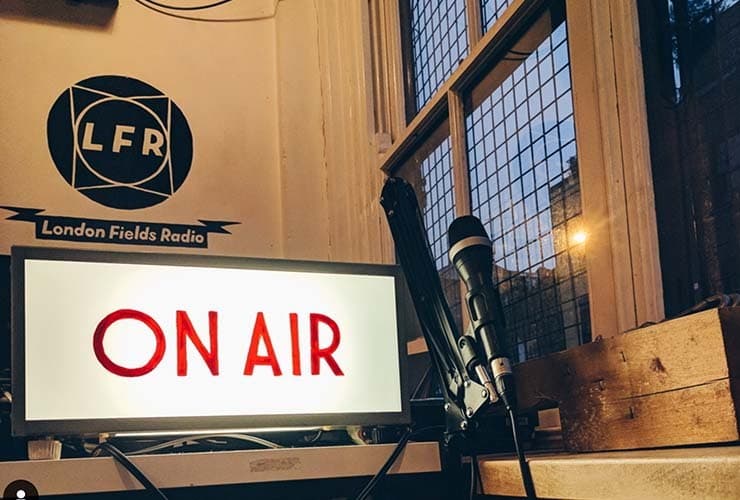 Image shows the London Fields Radio studio, featuring a microphone, their logo on the wall and a lit up 'on air' sign