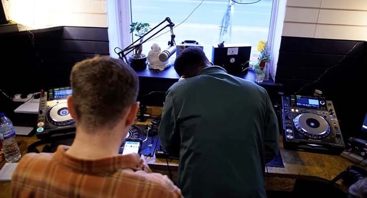 Image shows two young men in the radio studio at melodic distraction. One man is on his phone while the other is on the DJ decks.