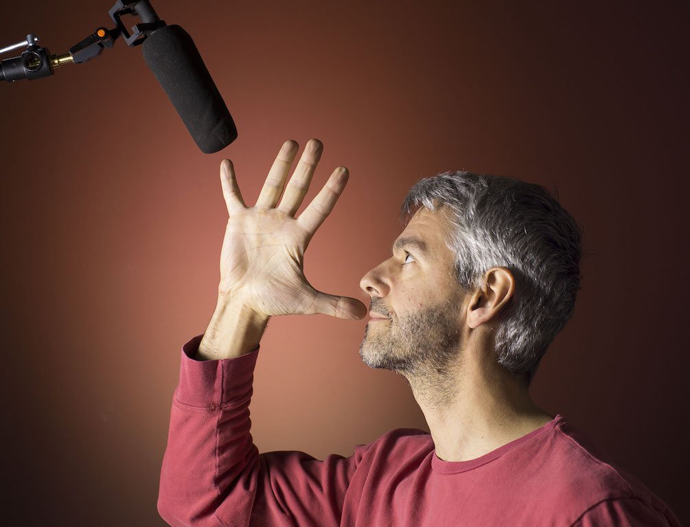 This is a chocolate brown backdrop. A white man with grey hair stands in the forefrnt. His hand is raised in a five to his face, with his thumb touching his lip as he stares forward. Above him is a black mic on a boom arm. He is wearing a red long sleeved t shirt.