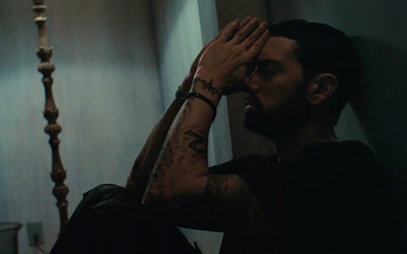 Image shows the rapper Eminem, sitting in a dark room against a wall with his hands on his head.