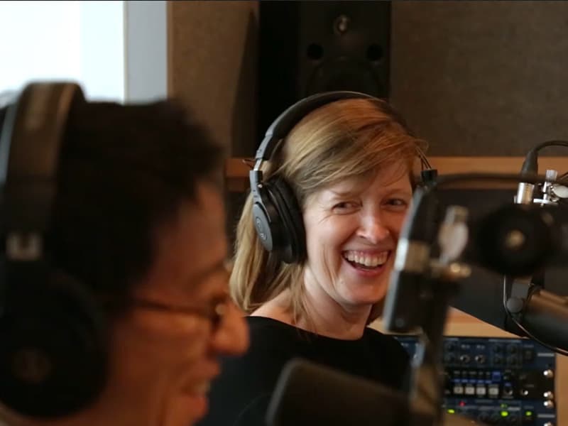 Image shows a female Monocle radio presenter in focus and laughing, and a male Monocle radio presenter out of focus and also laughing.