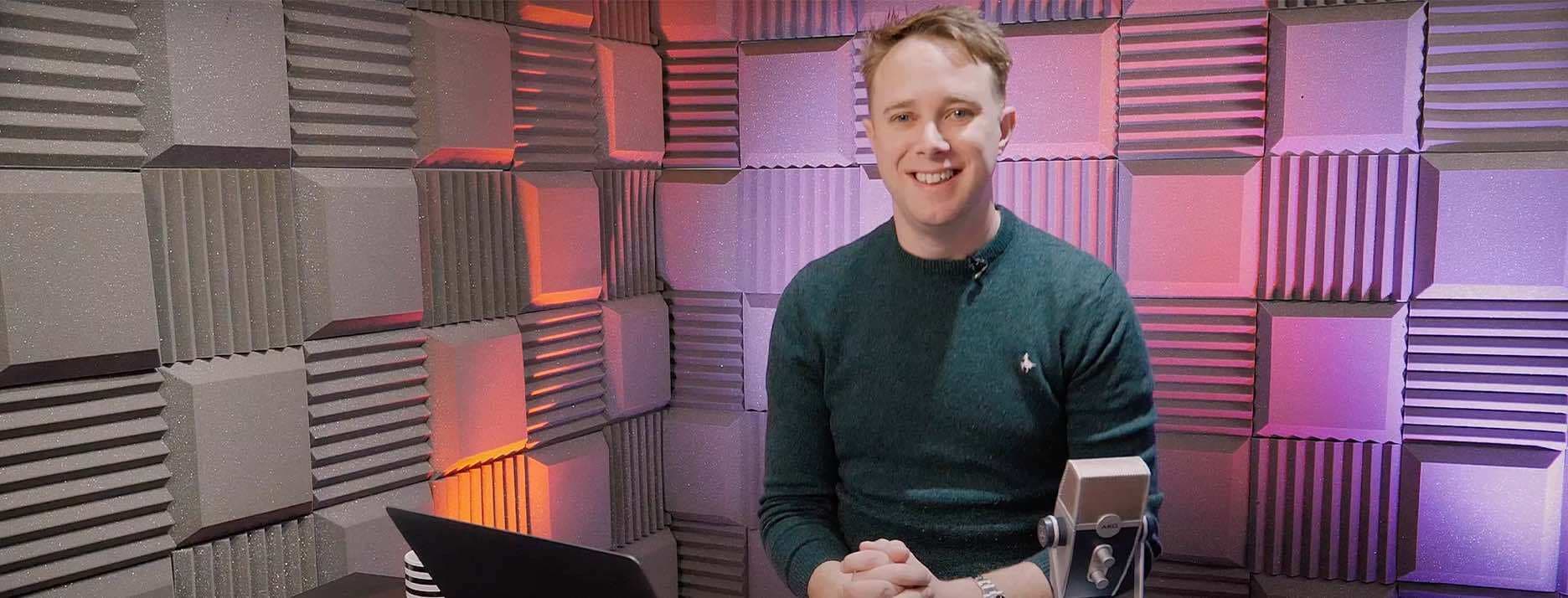 Image shows James Mulvany, a white man, sat at a desk with a laptop with foam tiles on the walls.
