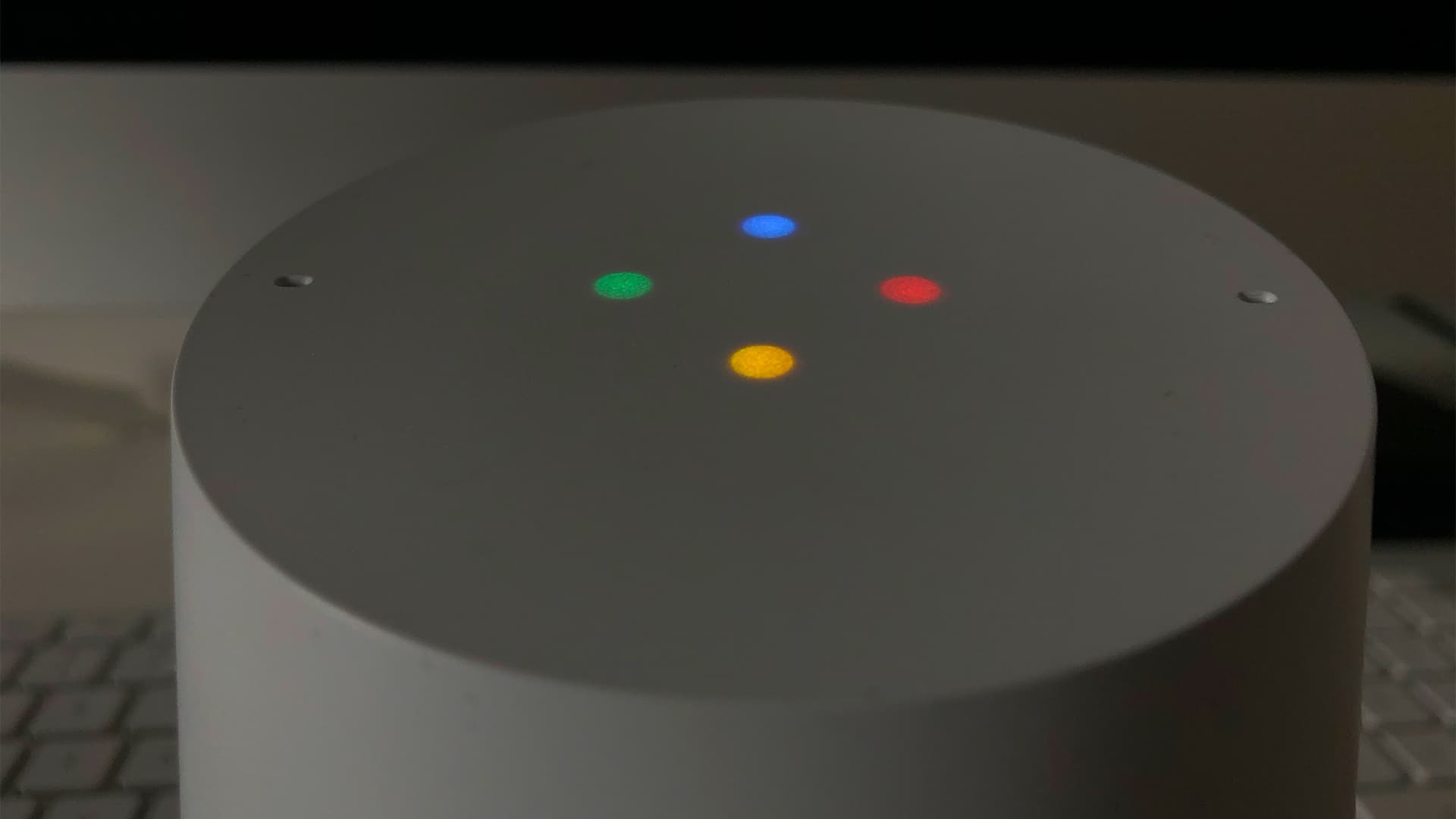 Image shows a the top of a grey Google Home smart speaker, zoomed in. All the display lights are on.