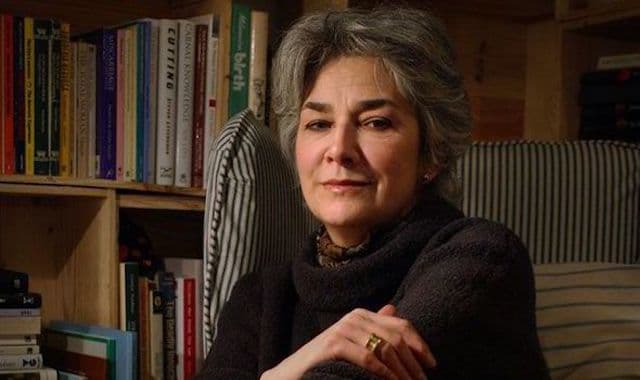 Image shows Anne Raeburn sat in an armchair in front of a bookcase.