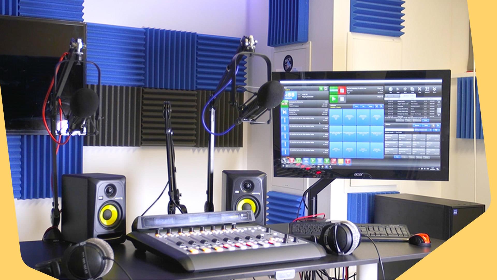 This is an example image of Radio Station Equipment For A Professional Studio Setup. There is a monitor on the wall on the right. On the main part of the desk is a mixing desk. The background shows blue and black soundpanels.
