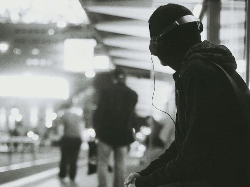 Image is a black and white photo of a person listening to headphones at a train station.