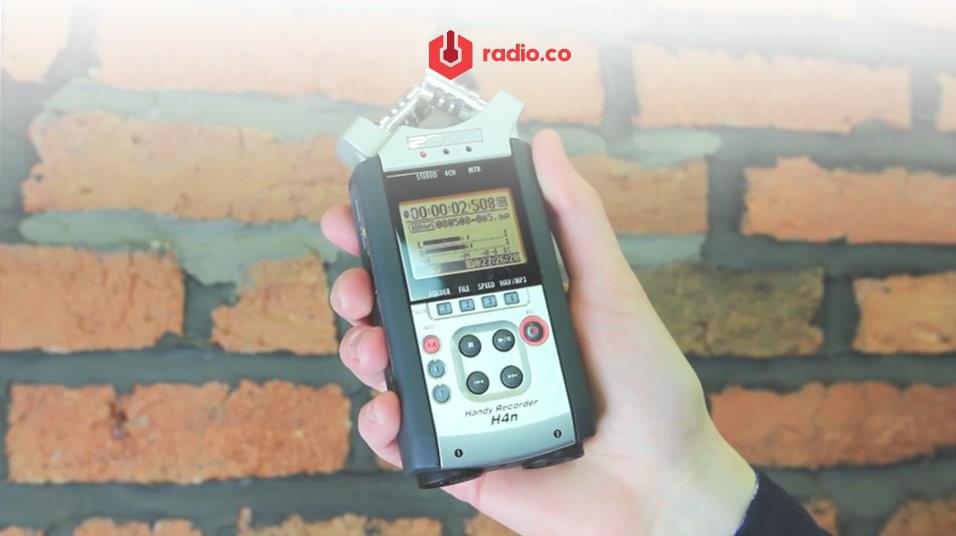 A white hand holds a rectangular microphone with an orange screen. The backdrop is a brick wall. At the top of the image is a red radio.co logo.