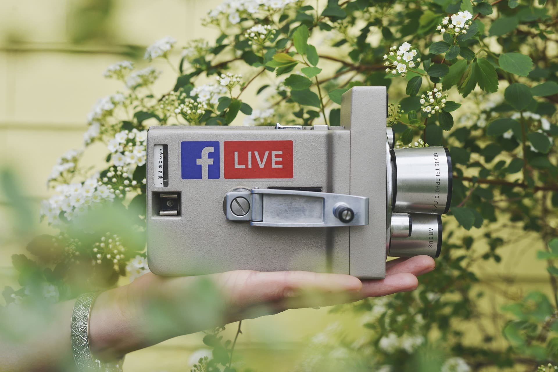Image is a hand in a garden, holding an old metal video camera that has a Facebook Live sticker on it.