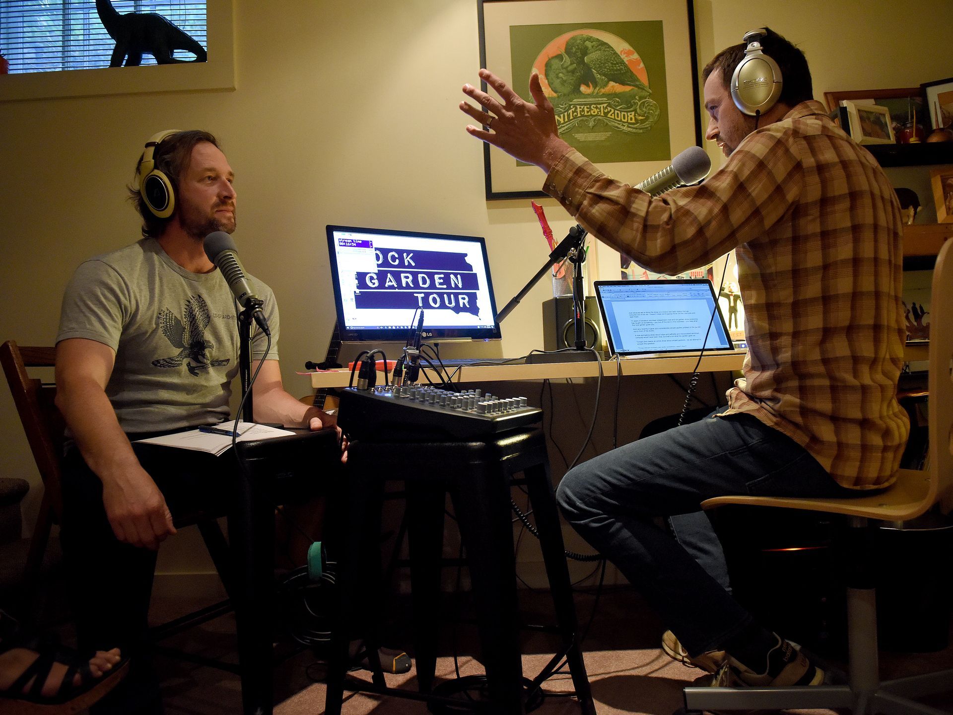 This is an image taken in the Rock Garden radio studio. A white man on the left has long dark hair and a short beard. He listens to silver headphones and speaks into a silver microphone. He is wearing a grey shirt. On the right isa man with a white and brown plaid shirt and silver headphones, presenting in an animated way to his coleague. He is stood in front of a silver mixing desk and a white and blue Rock Garden Tour monitor. The background is neutral but with a framed green and orange art print on the wall.