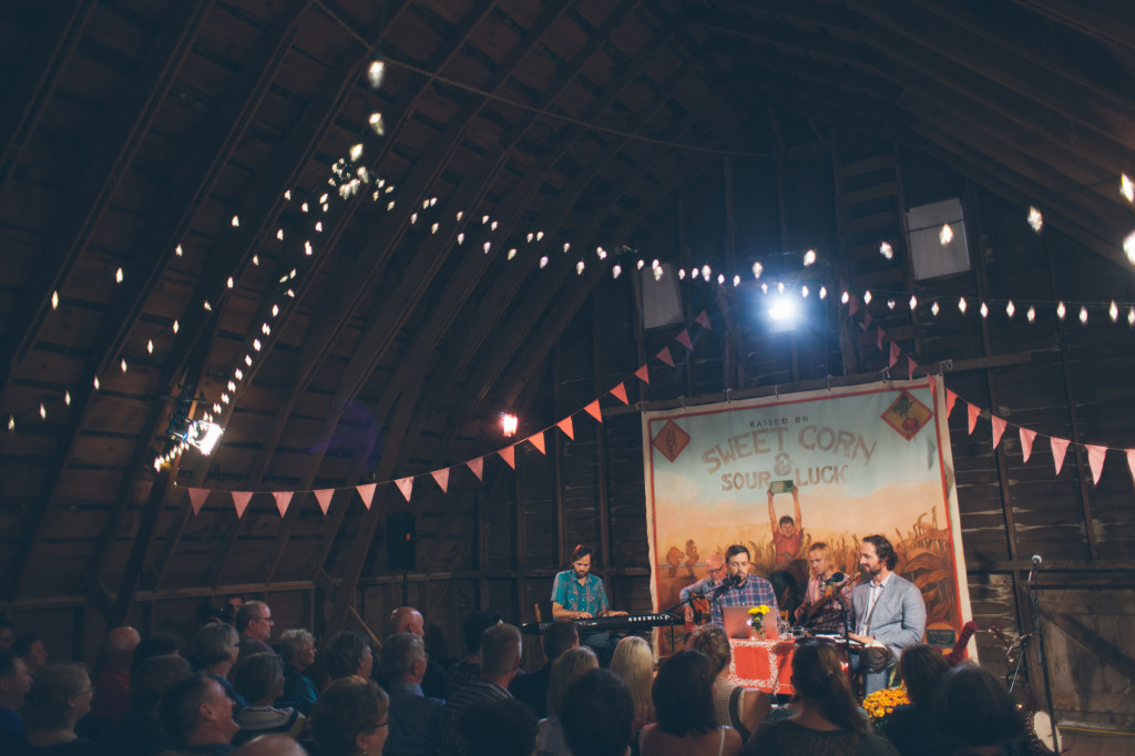 A large dark barn is pictured, with fairy lights and warm lighting strung across the walls, with red banners. There is a backdrop, illustrated, with Sweet Corn Sour Luck. There is a a band in front of it. The audience is full and we see the backs of their heads.Rock Garden Tour Radio Crowd