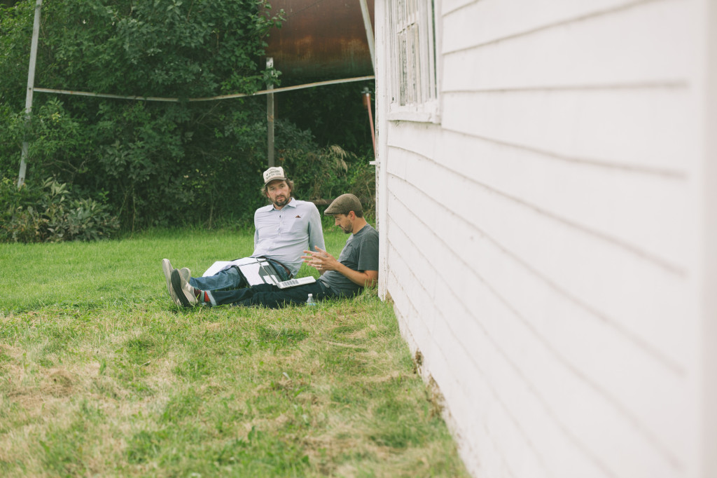 There are two white men, one in a white shirt with a white and black cap, and one with a grey hat and a grey shirt next to him. They are both sat on the grass next to a white wooden house. The men discuss something over a Macbook.
